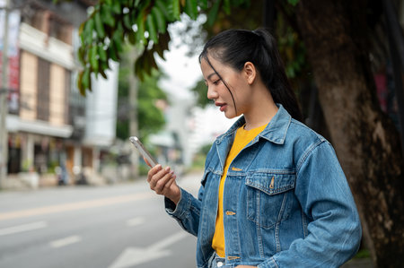 A side view of a beautiful, focused Asian woman in a denim jacket standing on a sidewalk, checking her smartphone as she books a taxi on an app or waits for someone.の写真素材