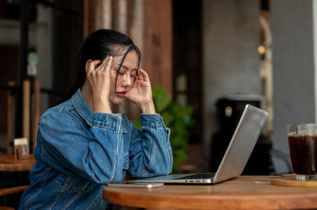 A side view of a stressed, thoughtful Asian woman in a denim jacket, feeling headache and tired, facing a problem with her project while working on her laptop remotely from a coffee shop.の写真素材