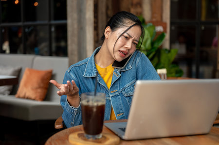 A stressed, busy, dissatisfied Asian woman in a denim jacket is on the phone with her colleague, complaining about a problem while working on her laptop remotely from a coffee shop.の写真素材