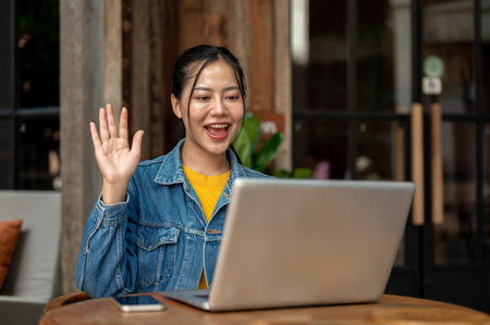 A beautiful, cheerful Asian woman in casual clothes is waving her hand during an online meeting on...