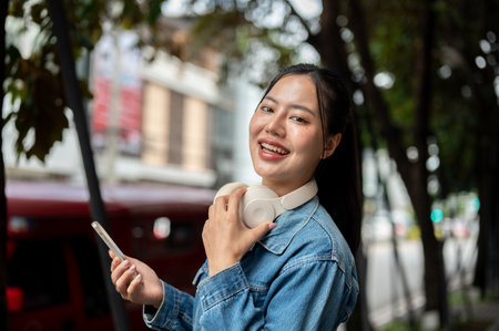 A beautiful, positive Asian woman in a denim jacket is standing on a sidewalk, holding a smartphone and smiling at the camera. waiting for someone or a taxiの写真素材