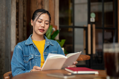 A charming, positive Asian woman in a denim jacket is reading a book while sitting in a coffee shop. people and lifestyle conceptsの写真素材