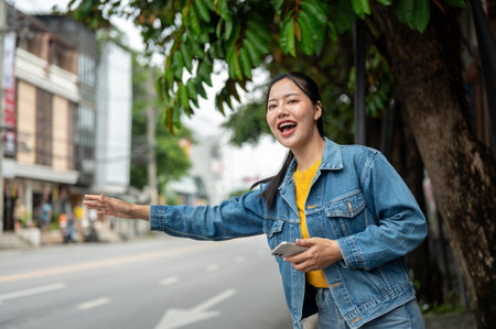 A beautiful, cheerful Asian woman in a denim jacket is standing on a sidewalk, hailing or catching a taxi in the city. people and public transportation conceptsの写真素材