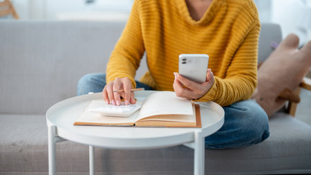 A close-up of a woman in a cozy sweater sitting on a sofa at home, using her phone and a calculator...