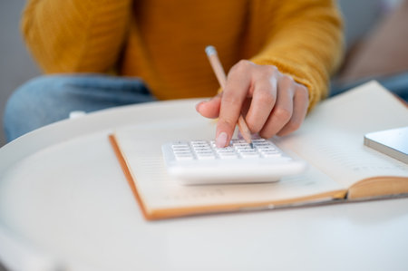 A close-up of a woman sitting on a sofa at home, using a calculator to manage her household expenses...