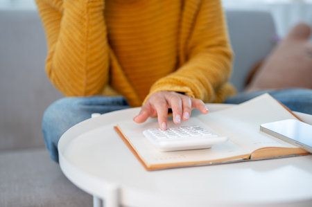 A close-up of a woman sitting on a sofa at home, using a calculator to manage her household expenses and calculate her monthly savings. people and financial conceptsの写真素材