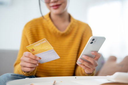 A close-up of a happy Asian woman in a cozy yellow sweater sitting on a couch and holding her smartphone and credit cards, shopping online at home. e-commerce, cashless payment, mobile bankingの写真素材