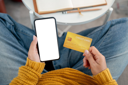 A top view of a woman in casual clothes holding a credit card and her smartphone on a sofa at home, shopping online or using a mobile banking app. with the smartphone displaying a white screen mockup.の写真素材