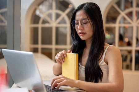An attractive Asian woman is focused on working on her laptop, typing on the laptop keyboard while...