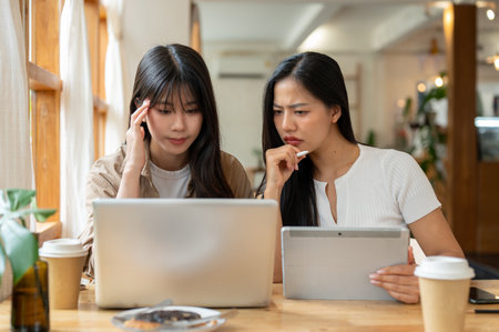 Two serious and stressed young Asian female friends are working remotely together from a coffee shop, focused on a laptop screen as they discuss work to solve problems.の写真素材