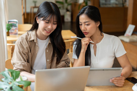 Two positive young Asian female friends are working remotely together from a coffee shop, discussing work while using a laptop and a digital tablet.の写真素材