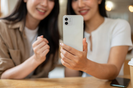 A close-up of two cheerful Asian females sitting in a coffee shop, enjoying a conversation while looking at something on a smartphone. chatting, gossip, girls day, lifestylesの写真素材