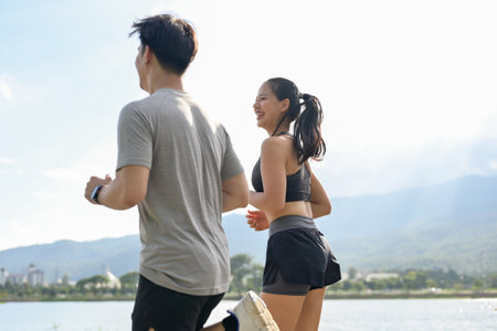 A rear view of a carefree and active Asian couple in sportswear enjoying a run or jog together by the lake on a sunny day. girlfriend and boyfriend, healthy lifestyle, workout, outdoor activityの写真素材