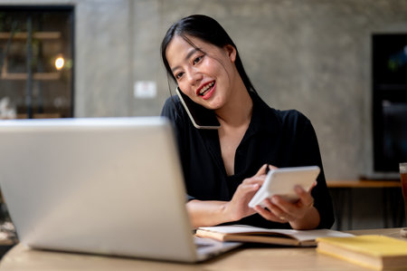 An attractive, busy Asian businesswoman is multitasking, talking on the phone with a client, focusing on her computer, and using a calculator while working at her desk in the office.の写真素材