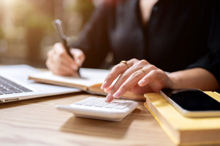 A close-up of a businesswoman's finger tapping on a calculator while taking notes in a book, working at a table indoors. financial, accounting, budget and expenses planning, taxの写真素材