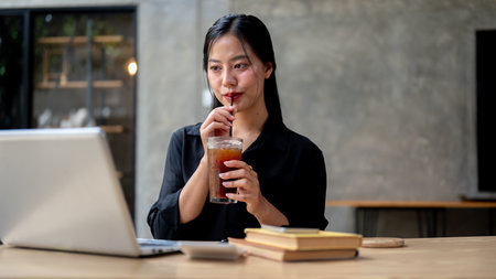 A beautiful Asian businesswoman enjoys iced coffee while working on her laptop, sitting at a table indoors and working remotely from a coffee shop. businesspeople, lifestyles, technologyの写真素材