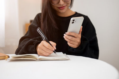 A close-up of a beautiful, positive woman in a black sweater writing in a book and using her smartphone while sitting at a table indoors. doing homework, taking notes, making listの写真素材