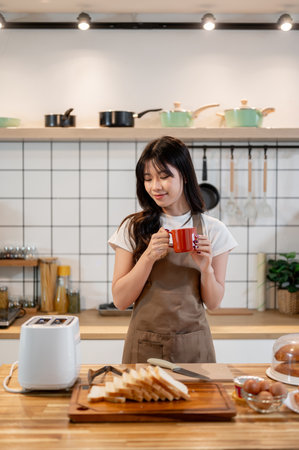A beautiful, positive Asian woman wearing an apron is enjoying coffee in the kitchen while preparing her breakfast. home cooking, domestic life, lifestylesの写真素材