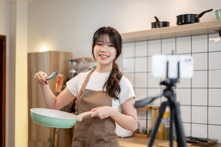A pretty and happy young Asian female food content creator or chef in an apron is recording a video while cooking in the kitchen, showing her food to the camera.の写真素材