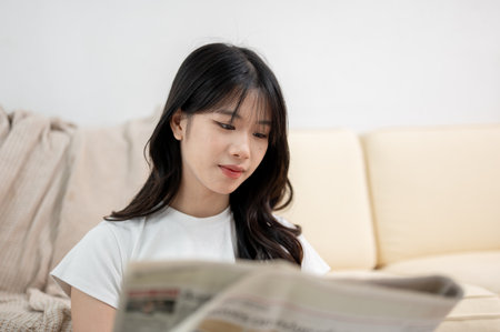A positive young Asian woman is focused on reading a newspaper in the living room in the morning....