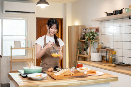 A pretty, happy young Asian woman is enjoying cooking in the kitchen, whisking eggs in a bowl while making breakfast. home cooking, domestic life, lifestyles, foodの写真素材