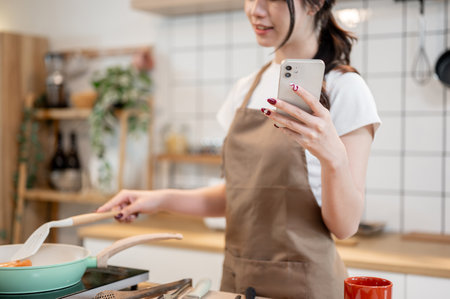 A close-up of a beautiful, happy Asian woman in an apron is holding her smartphone while cooking in the kitchen, checking messages on her phone while frying something in a pan.の写真素材