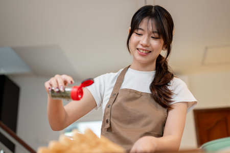 A beautiful, happy young Asian woman in an apron is sprinkling pepper on her food, enjoying cooking in the kitchen at home while preparing breakfast. the concept of people and foodの写真素材