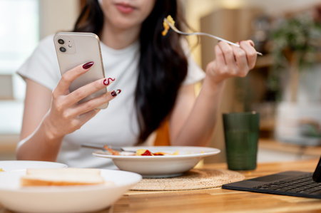 A close-up of a happy young Asian woman enjoying chatting or scrolling on her phone while eating breakfast at a dining table in her kitchen at home. image with a copy spaceの写真素材