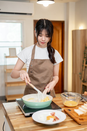 A charming, happy young Asian woman in an apron is cooking in the kitchen, making scrambled egg in a...