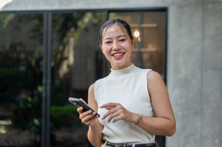 A portrait of a beautiful, cheerful Asian businesswoman holding a smartphone in her hand, standing...