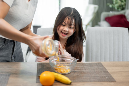 A young, adorable Asian girl is sitting at the dining table, excited to eat her cereal in the morning. A caring mom is pouring cereal from a jar for her daughter.の写真素材