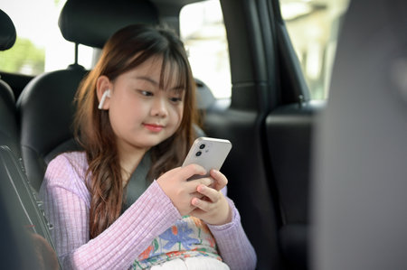 A young, adorable Asian girl is using her smartphone and listening to music through her earbuds while traveling with her parents, sitting in the backseat of a car with her seatbelt on.の写真素材