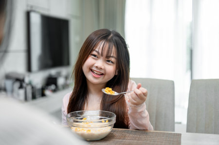 A young, adorable Asian girl is enjoying having breakfast with her mom at a dining table, eating a...