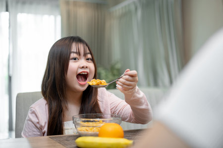 A young, adorable Asian girl is enjoying having breakfast with her mom at a dining table, eating a bowl of cereal. happy family moments, kids at home, foodの写真素材