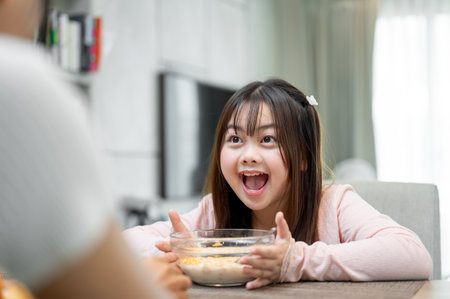 A young, adorable Asian girl is enjoying having breakfast with her mom at a dining table, eating a bowl of cereal. happy family moments, kids at home, foodの写真素材
