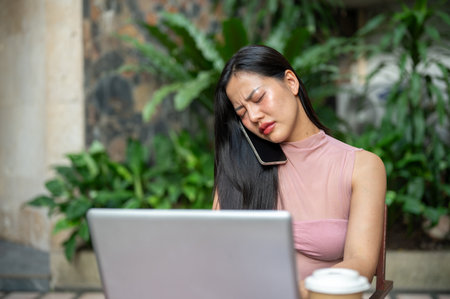 A stressed, thoughtful Asian businesswoman in casual clothes is having a serious phone conversation while working on her laptop remotely from a coffee shop, seated at an outdoor table.の写真素材