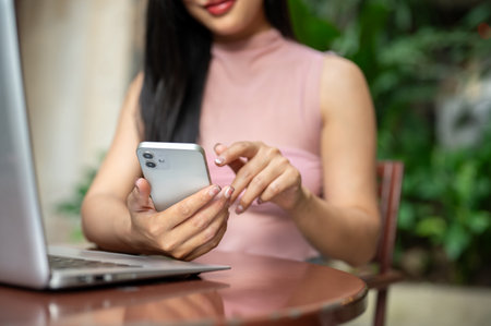 A close-up of a woman in casual clothes using her smartphone while sitting at an outdoor table of a cafe with her laptop, working remotely.の写真素材