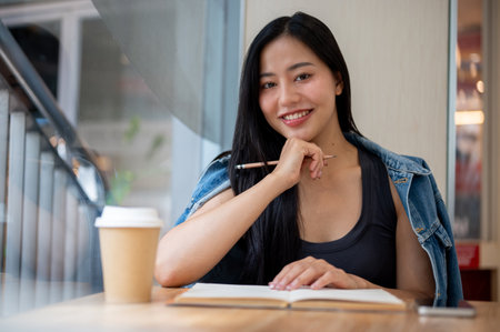 A portrait of an attractive Asian woman in casual clothes sitting by the window in a coffee shop with her diary and a coffee cup on the table. people and lifestyles conceptsの写真素材