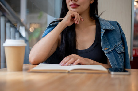 A close-up of a beautiful, thoughtful Asian woman sitting by the window in a coffee shop with her diary, contemplating something as she gazes outside.の写真素材