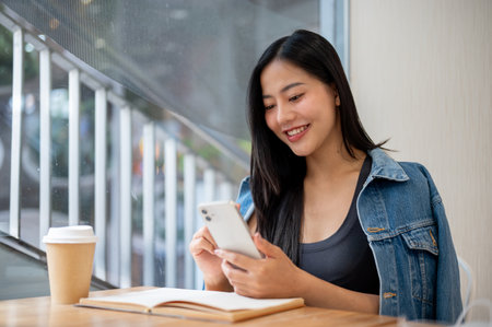 An attractive, smiling Asian woman in casual clothes is reading text messages on her smartphone while sitting in a coffee shop. people, wireless technology, lifestylesの写真素材