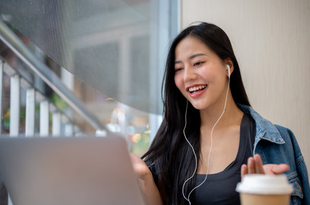 A smiling, charming Asian woman in casual clothes is having an online meeting with her team, wearing...