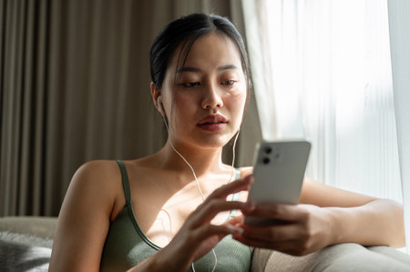 A beautiful Asian woman reading chat on her smartphone while listening to music through her earphones on a sofa in the living room. a cozy, warm atmosphere with sunlight streaming through the window.の写真素材