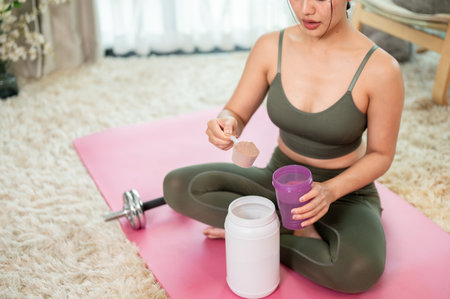 A close-up of a healthy, slim Asian woman in yoga clothing sitting on a yoga mat, preparing a protein drink after her workout at home. healthy lifestyle, wellbeing, nutrition, image with copy spaceの写真素材