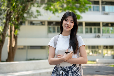 Portrait of a cute Asian female college student in casual clothes with a backpack, standing on her college campus, holding a book and smiling at the camera. people and education conceptsの写真素材