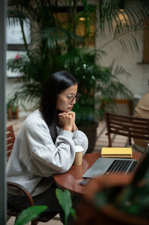A beautiful, focused Asian woman in a cozy sweater and eyeglasses works remotely from a coffee shop,...