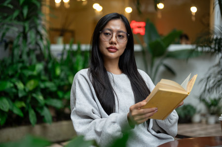 A calm, thoughtful Asian woman in a cozy sweater and eyeglasses reads at an outdoor table in a coffee shop, surrounded by green plants. people and lifestyles conceptsの写真素材