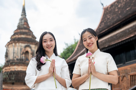 Two beautiful Asian women in traditional Northern Thai-Lanna dresses stand in a temple, holding pink lotuses and smiling at the camera. Thai culture, Asia traveling, Buddhist Holidaysの写真素材