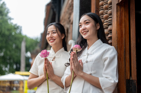 Two beautiful Asian women in traditional Thai-Lanna dresses stand in a temple, holding lotuses in a prayer posture and smiling away from camera. Thai culture, Buddhistの写真素材