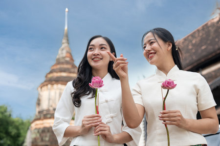 Two beautiful, smiling Asian women in traditional Northern Thai-Lanna dresses stand in a temple, holding pink lotuses and enjoying a conversation together. Buddhist, Thai cultureの写真素材