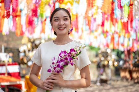 An attractive, smiling Asian woman in a traditional Northern Thai dress stands in a temple, holding a bouquet of orchids with colorful lanterns in the background. Loy Krathong festival, Thai cultureの写真素材